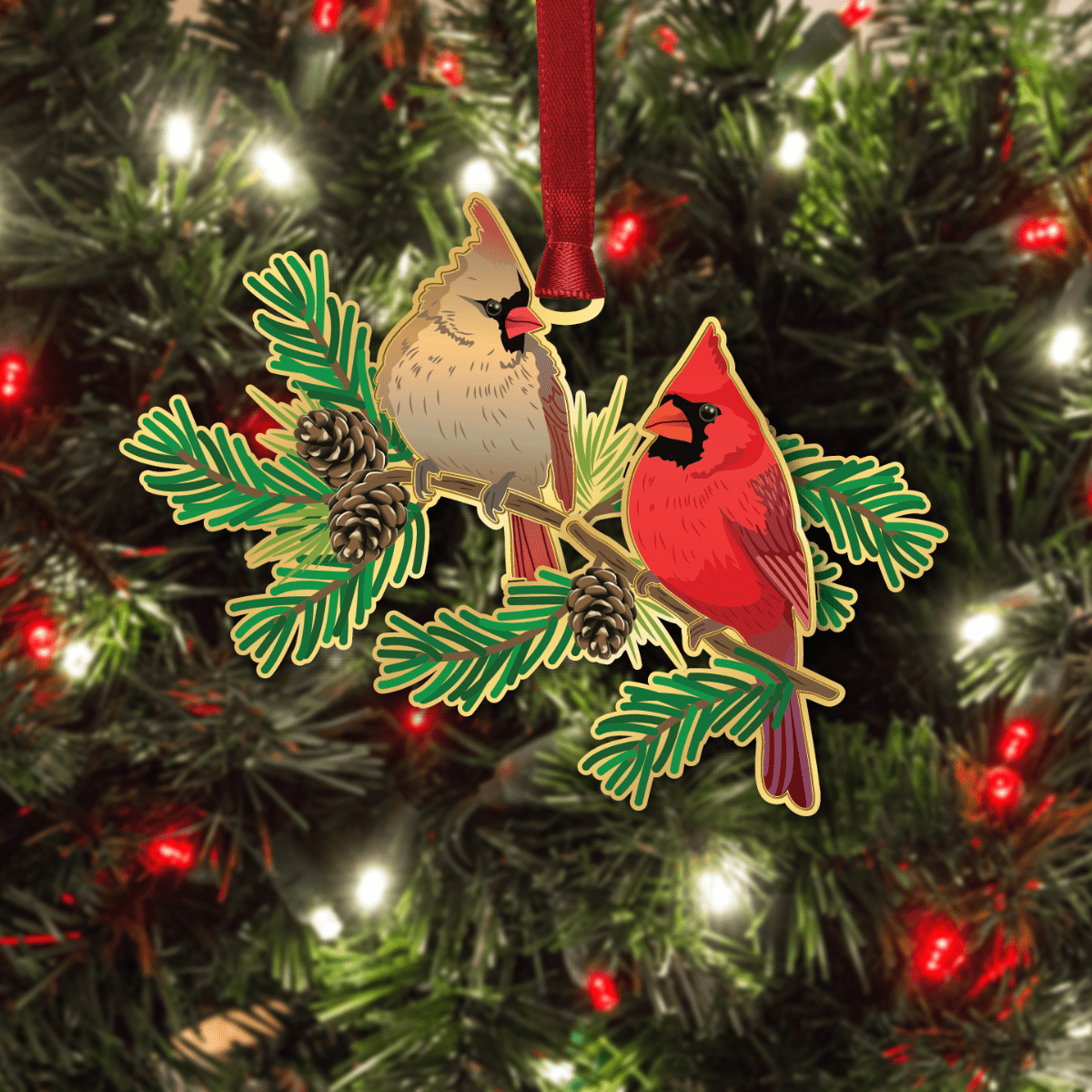 Ornament featuring a pair of northern cardinals perched on evergreen branches with pinecones, displayed on a decorated Christmas tree